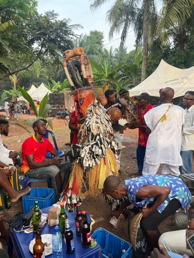 An outdoor cultural gathering in Nnobi, Nigeria, is captured, centered around an elaborately dressed masquerade figure. The masquerade dominates the foreground and midground, adorned with numerous bundles of what appear to be currency notes or paper strips, creating a textured, almost feathery covering. Its costume also features raffia or straw elements, red and orange fabrics, and an intricate, dark headpiece.
Many people are gathered around, observing the masquerade. In the immediate foreground, a man in a red t-shirt sits on a blue plastic chair, looking towards the activity. Nearby, a blue plastic crate serves as a makeshift table, laden with various beverage bottles (some alcoholic, some water) and stacks of money. Another man, wearing a blue floral shirt, bends over, handling money near a wicker basket. Other individuals in casual and traditional attire are visible standing and sitting throughout the scene.
In the background, lush tropical vegetation, including palm trees and banana plants, indicates a verdant, likely rural or semi-rural setting. White canopy tents are erected, providing shade. One tent clearly displays the text "DESIGNER FEES SERVICES". Speakers for a sound system are also visible in the mid-distance, suggesting an organized event. The ground is dirt. The time appears to be daytime under an overcast or bright sky, creating a vibrant yet diffused light. The overall atmosphere is that of a lively community celebration or traditional festival.