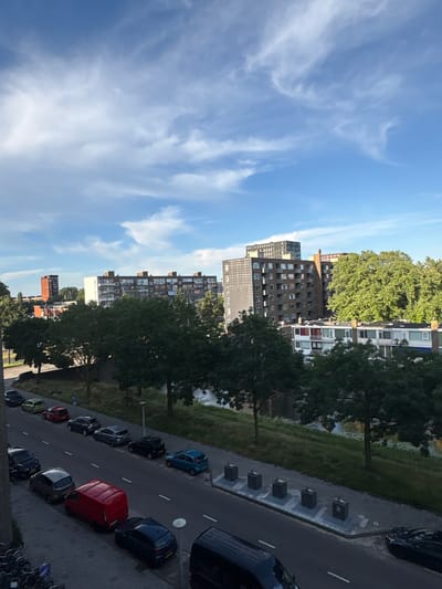The image shows a street in Amsterdam, Netherlands, with parked cars lining one side of the road. In the background, there are several apartment buildings and trees. The sky is blue with some white clouds. There are no people visible in the image. The scene appears to be a typical urban environment.