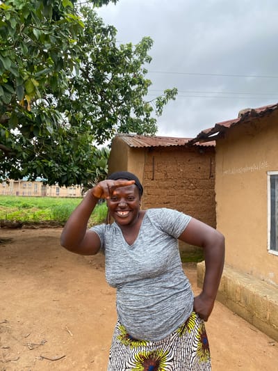 In the city of Riyom, Nigeria, a woman with dark skin and a black headscarf stands outdoors. She is smiling broadly and holding her right hand up to her forehead, creating a gesture. She wears a grey, V-neck t-shirt and a vibrant, patterned skirt featuring yellow, white, and black designs with blue and yellow circular elements. Behind her, a brown clay building with a corrugated metal roof is visible, showing some signs of wear and tear, with a window on the right side. White chalk writing is inscribed on the building's wall. To the left of the building, a large green tree with dense foliage shades the scene. In the background, a long, light-colored building with multiple windows and a tiled roof can be seen, suggesting a more institutional structure. The ground is composed of reddish-brown dirt, scattered with small twigs and dry leaves. The sky overhead is overcast and grey.