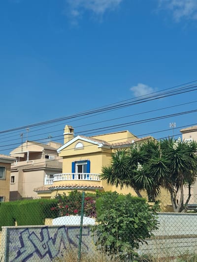 The image shows a residential area in Playa Flamenca, Spain, on a clear day. In the foreground, there is a chain-link fence with some graffiti on a concrete wall behind it. Dry grass and some green bushes are visible in front of the fence. Behind the fence, there are several houses with tiled roofs. The houses are painted in pastel colors like yellow and pinkish-beige. One yellow house has a distinctive blue shuttered window and a white balcony with railings. A large palm tree with white flowers is situated in front of the yellow house. In the background, overhead power lines crisscross the sky, which is a bright blue with a few scattered white clouds. The scene depicts a sunny day in a Mediterranean-style residential neighborhood.