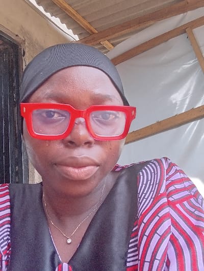 The scene depicts a close-up portrait of an adult female in Ayob, Nigeria. The individual has dark skin and wears a black head covering. She is seen wearing large, red, rectangular eyeglasses, with her eyes visible behind the lenses. Her mouth is closed, and her expression is neutral. She is dressed in a garment with a distinctive wavy pattern featuring red, purple, and black colors, partially overlaid by a black V-neck top. A thin, gold-colored necklace with a small, clear gemstone pendant is visible around her neck. The background indicates an outdoor or semi-sheltered environment, showing a dark doorway and a light-colored wall to the left, and wooden support beams, white tarp-like material, and corrugated metal roofing to the right.