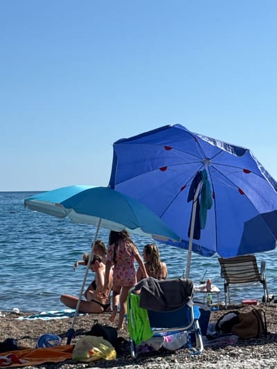 The image captures a sunny beach scene in Rafailovići, Montenegro, under a clear, cloudless blue sky that occupies the upper third of the frame. The horizon line is visible and flat. The sea, a deep blue, is calm with minor ripples extending to the pebble beach in the foreground. A small, dark, rectangular object floats on the water in the mid-distance on the left.
Two open beach umbrellas are prominent. A smaller, light blue umbrella with a white pole stands on the left. To its right, a larger, dark blue umbrella with a white pole features red accents on its underside ribs, and a dark blue and a light blue/teal garment are hanging from its pole.
Several individuals are present on the beach. Partially visible under the light blue umbrella on the left is a person with long, light-colored hair, seemingly sitting or kneeling near the water. To their right, a person with light brown hair tied back is seated or kneeling on the pebbles, facing away from the viewer, wearing a dark bikini bottom. A young person, likely a child, with long light brown hair and wearing a light-colored floral or patterned dress, stands upright with their back mostly to the camera, positioned between the two umbrellas. Another individual is partially obscured by the large blue umbrella on the far right.
Various beach equipment is scattered on the dark pebble beach. These include an orange towel, a dark blue cap, a blue plastic item (possibly a snorkel mask), a translucent yellow plastic bag containing an orange object, and a black backpack in the immediate foreground. A blue foldable beach chair with a grey towel draped over it is visible under the large blue umbrella. On the far right, a striped beach chair (white and dark stripes) with a metal frame is set up facing the water. A small, light-colored table with a red bottle and another container rests near the water's edge behind the chairs on the right.
