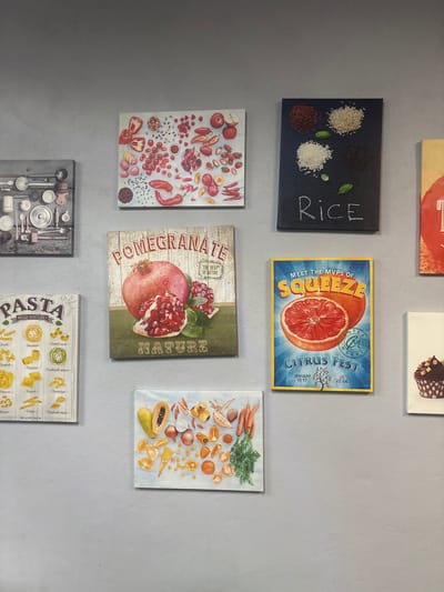 The image shows a collection of food-themed prints displayed on a light grey wall. The prints include images of rice, citrus fruits, pomegranates, an assortment of vegetables, and pasta, each with accompanying text descriptions or related words like "squeeze", "nature", and "what you like?" The image also features a kitchen utensil collage.