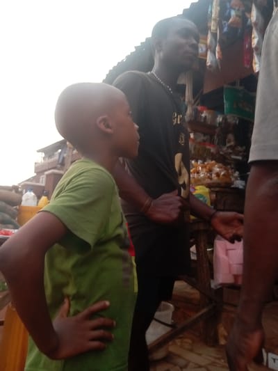 In Ogidi, Nigeria, a young boy with a shaved head stands in a green t-shirt, his hand on his hip, looking to the side. Behind him, a taller man with a black t-shirt and necklace stands with his hand extended, appearing to be offering or asking for something. They are in front of a wooden structure with various items displayed, including snacks and packages, suggesting a market or street vending setup. Another individual is partially visible on the right edge of the frame, obscuring the view. In the background, buildings can be seen, indicating an urban environment.
