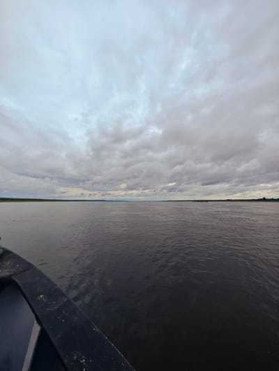 The image depicts a wide, calm river under a cloudy sky. The perspective is from the edge of a dark-colored boat, with a portion of its railing visible in the foreground. The river stretches out to the horizon, where a thin strip of green land, likely a shoreline with vegetation, is visible. In the distance, a dark, elongated object, possibly a barge or a long boat, is present on the water. The sky is filled with an overcast of grey and white clouds, suggesting a subdued or possibly rainy weather condition. The overall scene conveys a sense of travel or journey on a vast body of water. The location is identified as Zotino, Russia.
