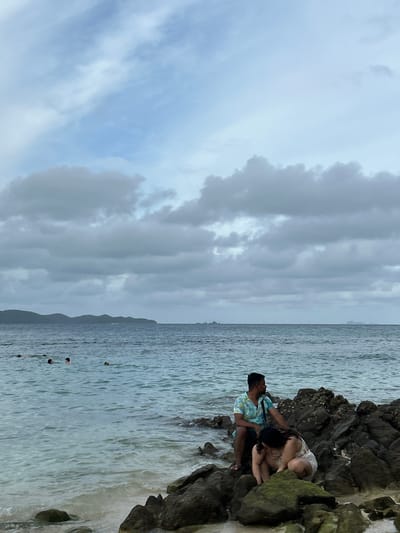 This is a full-shot photograph of a rocky beach at Tambon Phru Nai, Thailand, under a cloudy sky. In the foreground, two people are seated on dark, wet rocks near the shoreline. A man wearing a teal and white patterned short-sleeved shirt and dark shorts sits on the left, looking out towards the sea. A woman in a white outfit kneels on the right, facing downwards, seemingly examining something on the rocks. The sea is calm with gentle waves washing ashore. In the mid-ground, several people are visible swimming or wading in the water. The background features a distant, dark green, hilly island chain under a sky filled with scattered grey and white clouds.
