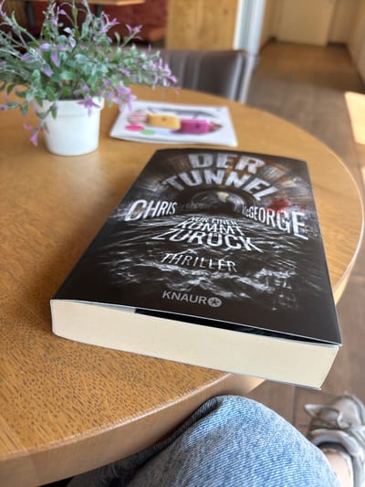 The image shows a book with a dark, dramatic cover titled "DER TUNNEL" lying on a wooden table. The title and author's name, "McGEORGE," are prominently displayed in large, white, distressed lettering. Below the author's name, the title continues with "NUR EINER KOMMT ZURÜCK" and "THRILLER." The cover art depicts a dark, vortex-like tunnel with streaks of light and shadow, suggesting a suspenseful or thrilling theme. The KNAUR logo is visible in the bottom right corner of the cover.
To the left of the book, a small potted plant with purple flowers adds a touch of color to the scene. A white card with colorful shapes is also partially visible behind the plant. To the right of the book, a person's legs clad in blue denim jeans and grey athletic shoes are visible, indicating someone is sitting at the table.
The lighting suggests it might be daytime, possibly indoors in a cafe or library, given the warm glow on the wooden table and the general ambiance. The overall impression is of a casual moment of reading or relaxation. The text on the book cover is in German.