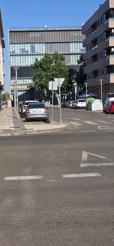 The image depicts a street scene in the city of Huesca, Spain, likely in a modern urban area. In the foreground, a paved street is visible with white lane markings and a pedestrian crossing. Several cars are parked along the sidewalk on the left side of the street, including a silver Volkswagen Passat with license plate "4750 CFT" in the immediate foreground. Behind it, other cars are parked, leading up to a sidewalk with streetlights.
On the right side of the street, across the pedestrian crossing, there are more parked cars, a green tent-like structure, and a white modular bin. In the background, a large, modern building with a glass and metal facade dominates the scene, flanked by another residential building with balconies. A large, leafy tree is situated between the street and the modern building. The sky is clear and blue, indicating a sunny day. There are no people visible in the image.
