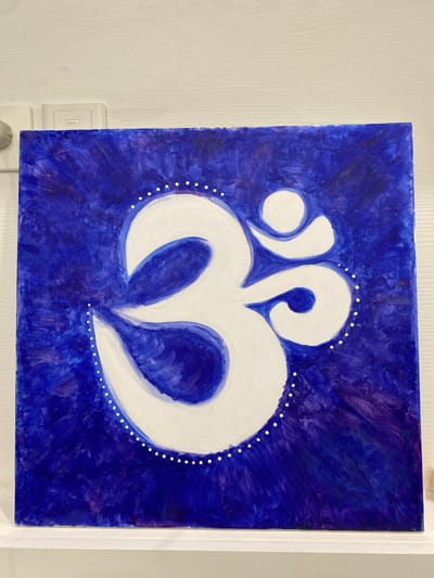 This is a photograph of a painting on canvas. The painting features a white abstract symbol against a deep blue background. The symbol is centrally located and has a symmetrical, organic form, resembling a stylized heart or a pair of abstract wings. It is outlined by a border of small white dots. The blue background is textured, with visible brushstrokes creating a sense of depth and movement. The painting is framed in white and hangs on a wall. To the left of the painting, a light switch with the brand name "Legrand" is visible. The wall appears to be white and textured, possibly wood paneling. The overall impression is one of contemporary art, possibly with spiritual or meditative connotations due to the symbol's form. There is no indication of time of day, weather, or specific emotions conveyed beyond the abstract nature of the artwork. The location context of Phú Quốc, Vietnam, does not provide any visual cues within the image itself.