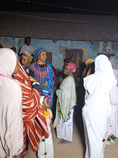 A group of people are gathered outside a building with a corrugated metal roof. Several women are wearing colorful headscarves and wraps, while others are wearing white hijabs. One young girl stands out with a red and white striped garment. A man stands in the background, partially obscured by the building. The overall atmosphere appears to be lively and communal.