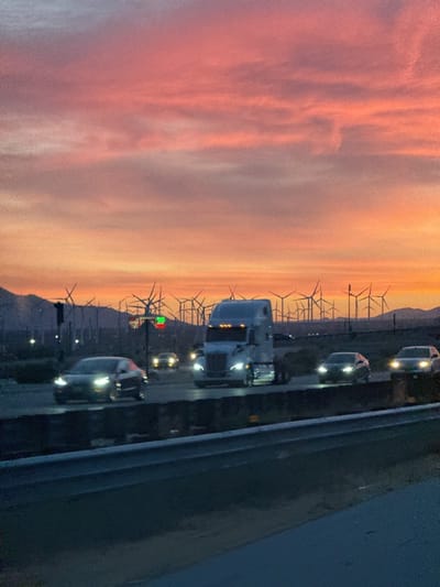 The image captures a sunset scene near Desert Hot Springs, United States, with a prominent array of wind turbines dotting the horizon. The sky displays a vivid gradient of pink, orange, and yellow hues. In the foreground, a highway is visible with several vehicles including a semi-truck and at least three cars with headlights on. A traffic light pole is on the left, with green lights illuminated. The road is separated by a grey concrete barrier. The overall scene suggests dusk or dawn near a highway and a wind farm outside of Desert Hot Springs.