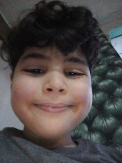 The image shows a close-up of a young boy's face. He is smiling slightly and looking directly at the camera. He has dark curly hair, brown eyes, and is wearing a grey t-shirt. The background is out of focus, but it appears to be a dark green leather-upholstered chair or couch. 

The provided coordinates (latitude: 31.227, longitude: 29.962) place the image in Alexandria, Egypt. However, the image itself does not provide any information about the location.  The scene is a simple, everyday portrait and does not suggest any specific location or activity.