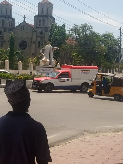 The image depicts a daytime street scene in Mombasa, Kenya, specifically at the Makadara/Mji Wa Kale ward near the Mkomani Roundabout. In the foreground, the back of an individual wearing a dark shirt and a patterned kufi cap is visible, facing away from the camera and towards the street.
The midground features an asphalt road. A red and white Kenya Posta pickup truck with an enclosed cargo bed, marked "POSTA Tracking Lines," is positioned on the left side of the road. To its right, a yellow auto-rickshaw (tuk-tuk) with a black canopy, displaying "TAXI 5 PASS," is also on the road. A person in a blue shirt is visible inside the tuk-tuk, presumably the driver. The road is bordered by a raised curb and a reddish-brown patterned sidewalk on the right.
In the background, a large, white monument shaped like a traditional Arabic coffee pot (dallah) stands on a circular pedestal. The pedestal bears partially visible text, "COUNTY GOVERNMENT OF MOMBASA." Behind this monument, a grand, light-colored stone church, identified as the Holy Ghost Cathedral, dominates the skyline with its two prominent bell towers, conical roofs, and a central cross. A large circular window is visible on the church's facade. Green trees are interspersed throughout the background, partially obscuring parts of the church and surrounding area. Overhead utility lines are visible against the clear sky.