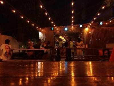 The image captures a vibrant night scene at a casual outdoor or semi-enclosed venue in Maputo, Mozambique. The perspective is low, looking across a highly reflective wooden tabletop in the foreground that shimmers with the warm glow of overhead string lights.

Numerous individuals are gathered, socializing and enjoying the evening. On the left, a person is seated with their back to the camera, wearing a white t-shirt with a prominent portrait of musician Chris Brown printed on it. They are at a wooden table with other patrons, with drinks and small items visible on the surface. Toward the center and right, more people are standing and mingling near what appears to be a bar or serving area. Two women are clearly visible there, one in a light green outfit and another in a light-colored top and pants.

The setting is illuminated primarily by strings of warm-toned bulb lights strung across the area, creating a festive and inviting ambiance. Additionally, a few blue and white disco-style lights add a dynamic element to the overhead lighting. The venue features a dark, possibly tarp-covered roof structure and a rustic-looking bamboo-slat wall on the right side. The overall atmosphere is relaxed and social, indicative of a night out at a bar or restaurant. The time of day is clearly night, and no other visible text is discernible in the scene apart from the portrait on the t-shirt.