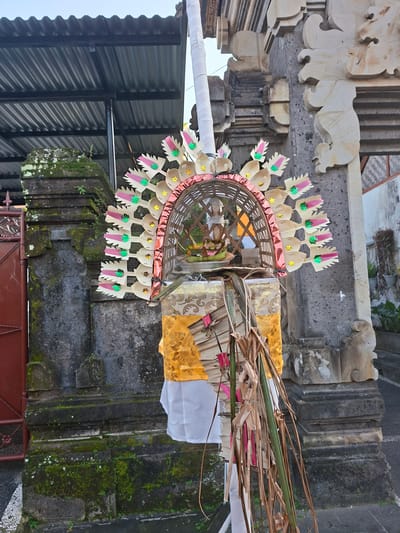 The image shows a decorative offering, known as a "Penjor," standing vertically against a stone structure in Blahbatuh, Indonesia. The Penjor is adorned with colorful paper cutouts, metallic accents, and palm leaves. It is draped with white and yellow fabric. The stone structure appears to be part of a temple or traditional Balinese building, with intricate carvings and moss growth visible. In the background, a corrugated metal roof and a red gate can be seen. The lighting suggests it is daytime, with natural light illuminating the scene. There are no people or animals visible in the image.