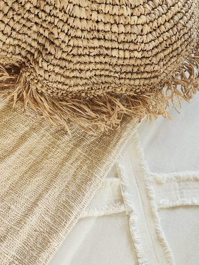 This media file is a close-up, overhead shot of woven natural materials. The main subject on the left is a textured object made of tightly woven straw or raffia, with a fringe of loose straw strands along its edge. This object appears to be a decorative item or part of a larger piece, like a basket or hat.
To the right of the straw object, there are two layers of fabric. The bottom layer is a coarse, loosely woven natural fabric in a light beige color, showing a clear woven texture. Overlapping this is a lighter, off-white fabric with a more uniform weave, characterized by distinctive horizontal bands of frayed threads along its edges. The arrangement suggests a still life composition, likely staged for a photograph.
The lighting is soft and diffused, indicating it could be natural light from a window or a studio setting. The overall aesthetic is organic, rustic, and minimalist, with a focus on texture and natural materials. There are no people visible, nor any specific clues about the time of day, weather, or precise location beyond the general context of Maitland, USA. The composition is abstract due to the close-up perspective, with no visible text. The scene evokes a sense of calm and natural beauty.
