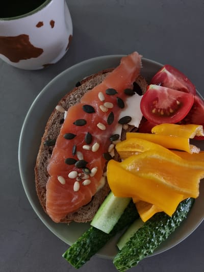 An overhead shot shows a plate of food on a grey surface, next to a cow-patterned pitcher. The plate contains a slice of dark bread topped with smoked salmon, white cheese, pumpkin seeds, and pine nuts. The salmon is pink and appears to be thinly sliced. Also on the plate are several wedges of tomato, slices of yellow bell pepper, and pieces of cucumber. The pitcher is white with brown splotches, resembling a cow's markings, and has a green rim. The food is arranged in a visually appealing manner, suggesting a meal or snack. The background is a plain grey surface, which could be a tabletop. No individuals are visible in the image, and no specific location in Ramat Gan, Israel, can be determined from the visual information provided.