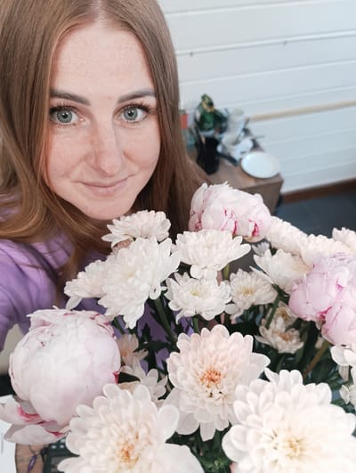 A close-up, medium shot shows a young woman with auburn hair and fair skin, dotted with freckles, looking into the camera. She is wearing a purple long-sleeved shirt and holding a bouquet of white and light pink peonies and chrysanthemums. The flowers are in the foreground, obscuring part of her face. In the background, a shelf holds various items, including a green figurine, stacked white dishes, and a white plate. The wall behind the shelf is made of horizontal wooden planks, painted white. The scene appears to be indoors. This image was taken in Chernogolovka, Russia.