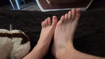 The image shows a close-up view of two feet resting on a dark brown, plush blanket. The foot on the left appears to be a female foot, with neatly trimmed toenails, some painted with French tips. The foot on the right is larger and shows more hair, suggesting it belongs to a male. The feet are positioned side-by-side, with the toes pointing upwards and slightly to the left. To the left of the feet is a portion of a fluffy stuffed animal, white with brown patches. In the background, out of focus, are hints of furniture and flooring. The scene is indoors, likely in a living room or bedroom. There is no discernible information in the image to indicate a specific location in Dobrich, Bulgaria.