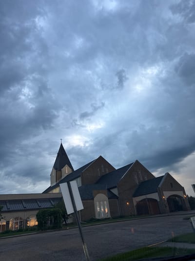 A church building with a tall steeple and a brick facade is shown under a cloudy, grey sky. The church appears to be a modern structure with large windows. In the foreground, there is a street with some puddles, suggesting recent rain. A blank white sign on a pole is visible to the right of the church. The image was taken in Stony Plain, Canada.