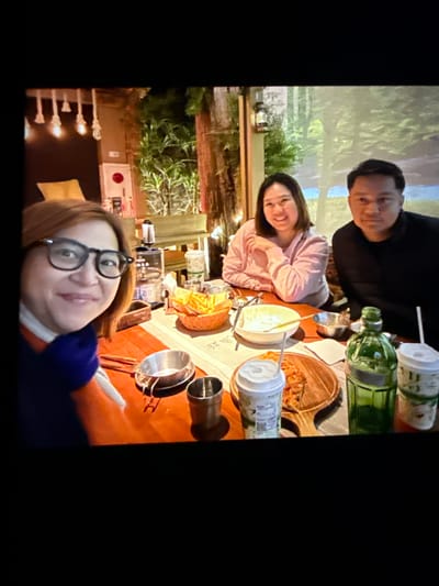 The image shows four people seated around a table, likely at a restaurant, enjoying a meal. The setting appears to be dimly lit, with a warm ambiance, possibly from string lights and lamps visible in the background. There are various food items and drinks on the table, including what appears to be pizza, fries, and beverages in cups and bottles.

The individuals are casually dressed, suggesting a relaxed social gathering. Two women and two men are present. The woman in the foreground, on the left, is wearing glasses and a scarf. The woman next to her is smiling, and the two men are seated further back.

The background hints at an outdoor or nature-themed setting, with greenery and what looks like a projected image of trees on a wall or screen. While the exact location in Taipei, Taiwan, cannot be definitively identified from the visual cues alone, the general atmosphere suggests a trendy eatery or a casual dining spot. The presence of multiple people enjoying food and drinks implies a convivial social occasion.