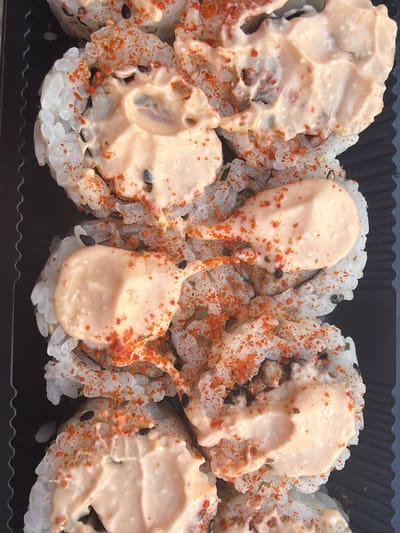 This is a close-up, top-down, eye-level shot of several pieces of sushi arranged in a black plastic container. The sushi appears to be a maki roll cut into bite-sized pieces, covered in a creamy, light orange sauce, and sprinkled with red chili flakes and black sesame seeds. The rice is white and appears to be sticky. The background is dark, showing the corrugated texture of the plastic container. There are no people or discernible landmarks indicating a specific location within Torrevieja, Spain. The image focuses exclusively on the food item.