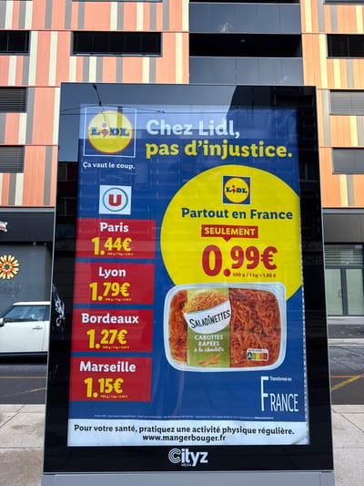 The image displays an outdoor urban scene at 30 Rue André Pingat in Reims, France. A large, vertically oriented, illuminated digital billboard occupies the center, showcasing an advertisement for Lidl.
The billboard's content is set against a blue background. On the right, a prominent yellow circle features the Lidl logo and the text "Partout en France SEULEMENT 0.99€" for a 500g portion (equating to 1.98€/kg). Below this, a clear plastic container of "Saladinettes Carottes Râpées à la ciboulette" (Grated carrots with chives) is shown, displaying a Nutri-Score 'A' label. The left side of the billboard presents comparative pricing from the "U" supermarket chain for the same 500g product in various French cities: Paris (1.44€), Lyon (1.79€), Bordeaux (1.27€), and Marseille (1.15€). The top section of the billboard includes the Lidl logo and the French slogans "Chez Lidl, pas d'injustice" (At Lidl, no injustice) and "Ça vaut le coup" (It's worth it). The bottom edge of the advertisement includes "Transformé en FRANCE" (Processed in FRANCE), a health recommendation "Pour votre santé, pratiquez une activité physique régulière. www.mangerbouger.fr" (For your health, practice regular physical activity), and the "Cityz Media" logo.
In the background, a modern multi-story building stands directly behind the billboard. Its facade is characterized by alternating vertical panels of orange and a lighter beige or light brown hue, separated by dark gray vertical strips. The building incorporates numerous dark-framed windows equipped with black horizontal louvers. To the left of the billboard, at street level, a dark storefront is visible, featuring a decorative, multi-petaled, orange and yellow floral emblem. A white car, identifiable as potentially a Mini Cooper, is partially visible parked on the pavement adjacent to this storefront. The ground surface is dark asphalt or concrete. No individuals or active interactions are present in the depicted scene.