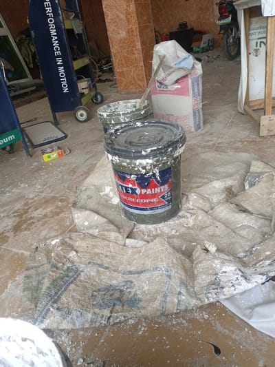 This image depicts a construction or renovation site in Hotoro North, Nigeria. In the foreground, a large, open paint bucket is visible, splattered with white paint and bearing the label "TEX PAINTS SCREEDING 20 LITRE". To its right, a torn, beige tarp is spread out, also covered in white paint splatters. Behind the tarp, a large bag of "DANGOTE CEMENT" is partially visible, with a wooden structure holding a white cloth to its right. Further back, a dark-colored vehicle, possibly a motorcycle, is parked. A blue cart with the words "PERFORMANCE IN MOTION" written vertically on its side is on the left. The ground is dusty and littered with debris, including scattered paint and small cardboard boxes. The overall environment appears to be an outdoor or semi-outdoor workspace, possibly a workshop or a storage area.