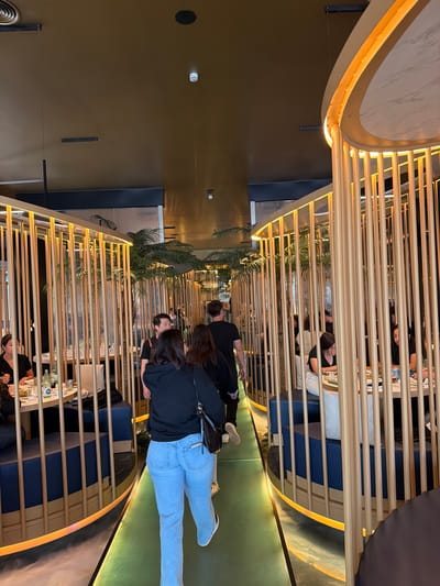 The image captures an interior shot of a restaurant in Torreforta, Spain. The scene shows a walkway bordered by curved booths. The booths are constructed with blue seating and divided by light-colored wooden slats that create a visual barrier. Patrons are seated in some of the booths, engaged in what appears to be dining. A woman wearing a black shirt and light blue jeans is walking along the green walkway. Another man walks in front of her, wearing a black shirt. Potted plants add a touch of greenery to the space. Overhead, the restaurant features an architectural design with curved elements and warm lighting. The walls are a solid brown color.