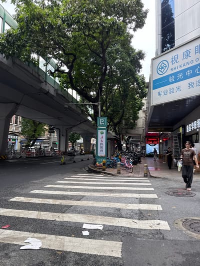 The image captures a street scene in Yuexiu District, China, with a pedestrian crosswalk prominently in the foreground. An elevated roadway or overpass dominates the upper portion of the frame, casting a shadow over the street. To the right, a building facade features a large sign with Chinese characters and the text "SHI KANG O". A woman walks across the crosswalk from right to left, carrying plastic bags. To the left of the crosswalk, parked bicycles are visible on the sidewalk, and further down the street, more businesses and a traffic light can be seen. The environment appears to be an urban setting with moderate pedestrian and vehicular activity.