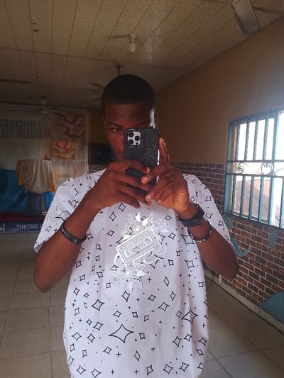 A male individual with short dark hair is depicted indoors, holding a black smartphone with a textured case in both hands, obscuring his eyes and nose, indicating he is taking a selfie. He wears a white short-sleeved shirt patterned with black geometric shapes and a central graphic of a cassette tape surrounded by flowers. A black wristband is on his left wrist, and a black watch along with a beaded bracelet are on his right wrist. The background features a large banner on the left partially displaying "THRONE OF MERCY CHAPEL" and imagery of baked goods, positioned behind a table draped with light-colored and blue fabric. To the right, a window with blue metal security bars and a section of a brick-patterned wall are visible. The ceiling consists of light-colored panels with exposed light fixtures. A light-colored floor and a partial blue seating area are also present. The scene is located within a religious establishment, specifically a chapel, in Mgbuoshimini, Nigeria.