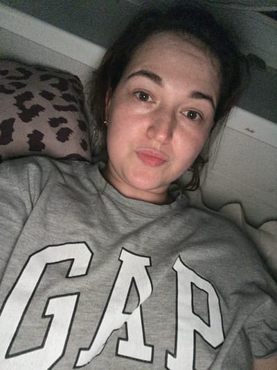 This is a close-up, eye-level selfie of a young woman lying down. She has fair skin, brown hair, and is wearing a grey Gap sweatshirt. The background appears to be a bed with a leopard-print pillow. The image has a grainy quality, suggesting it was taken in low light conditions. There is no discernible location in Chelyabinsk, Russia.