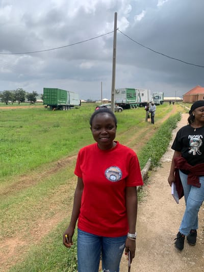 In Gwafan, Nigeria, two women stand on a dirt path in front of a row of shipping containers on trailers, under a cloudy sky. The woman in the foreground, wearing a red t-shirt and blue jeans, smiles at the camera. She holds a pen in her right hand and wears a watch and bracelet on her left wrist. To her right, a second woman, wearing a black t-shirt with a NASA logo featuring a cat astronaut, jeans, and a black beanie, looks to the right. She holds a book in her left hand. In the background, several green shipping containers are visible, some on trailers pulled by trucks. A few other people are walking further down the path. The environment is a grassy area with scattered trees and some buildings in the distance.