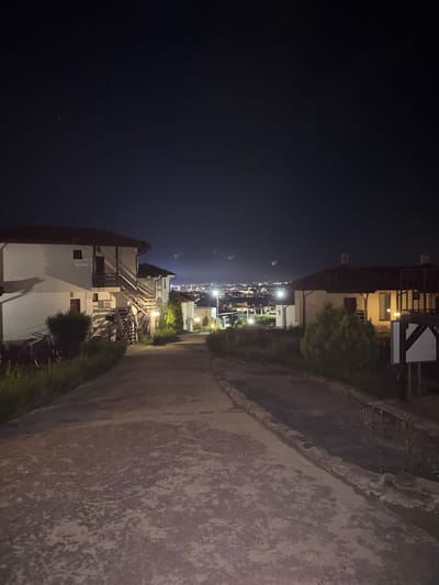 The image depicts a nighttime scene in Kosharitsa, Bulgaria, looking down a sloped, unpaved or worn asphalt road towards a distant illuminated city. The sky is dark with a few visible stars in the upper left quadrant.

In the foreground and midground, a rough-textured path, possibly concrete or asphalt, descends gradually, flanked by vegetation. On the left side of the path, a multi-story building, likely a guesthouse or villa, with white walls and a dark sloped roof, is prominently featured. It has multiple windows, external wooden staircases leading to upper-level balconies, and is illuminated by warm artificial light sources. Sparse green bushes and unkempt foliage are present along the left edge of the path.

On the right side of the path, a similar light-colored building with a dark roof is partially visible. A small, conical green shrub stands in front of this building. Further right, a wooden structure, possibly a signpost, is partially visible. Additional streetlights or external building lights are scattered along the path and near the structures, casting shadows and illuminating localized areas.

In the far background, a sprawling cityscape is visible, characterized by a dense concentration of artificial lights, indicating a populated area situated at a lower elevation than the viewer's position. There are no individuals, explicit actions, or interactions discernible in the frame.