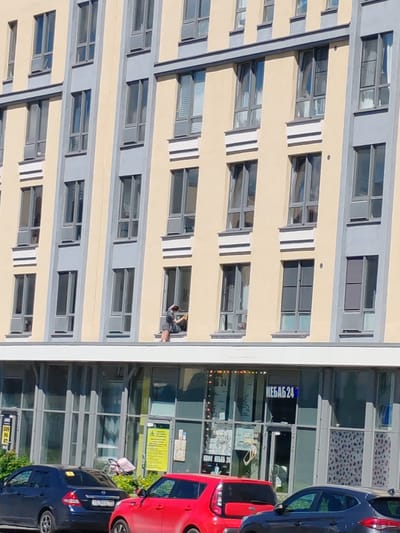 A person is sitting on the ledge of a tall building in Shushary, Russia, cleaning a window. The person is wearing dark clothing and appears to be using a yellow cleaning cloth. The building has a light yellow facade with grey window frames. Below the residential floors, there are commercial establishments with large glass windows. Cars are parked in front of these establishments, including a blue sedan and a red car. The sky is clear and the scene appears to be during daylight.