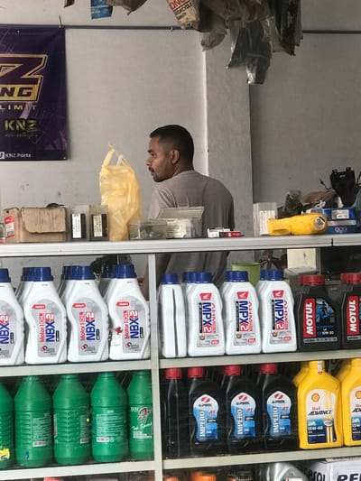 The image shows the interior of a shop in Rambot, Indonesia, likely specializing in motorcycle parts and accessories. A man, possibly the shopkeeper, is seen standing behind a shelf stocked with various brands and types of motorcycle oil, including Motul, Shell Advance, Impero, AHM Oil MPX2, and NBX-2. The shelf is compartmentalized, separating the different brands and oil types. The man is positioned next to the shelf, near an assortment of packaged motorcycle parts and a yellow protective cover. A banner with "KNZ LIMIT" branding is visible in the foreground. The overall environment appears to be a typical small retail shop.