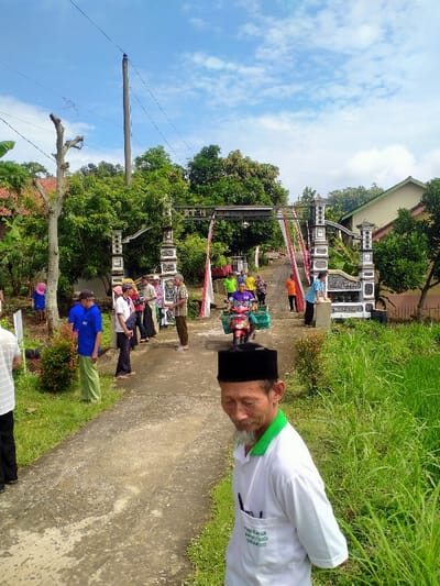 The scene takes place in Kecamatan Sambirejo, Indonesia, at latitude: -7.521, longitude: 111.094.  The location features a paved road with a decorative archway made of white and black pillars with a white metal roof.  Red and white fabric banners hang from the archway,  a motorcycle is parked in the middle of the road, and several people are standing around the archway.  There is lush vegetation on either side of the road, with some foliage in the background.  A person wearing a white shirt and green collar stands in the foreground, looking down at the ground.  Another person, wearing a blue shirt and a dark colored hat, is in the background.  Several other people are standing around the archway.  The sky is blue with white clouds.