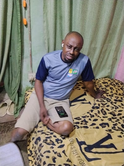 The image depicts an indoor scene in the city of Gwagwarwa, Nigeria. A bald adult male, appearing to be of African descent, with light brown skin, is seated on a bed. He wears a grey short-sleeved t-shirt with dark blue sleeves, featuring a multi-colored logo on the chest, and beige shorts. A white earbud is visible in his right ear. His facial expression displays a frown or grimace, with furrowed eyebrows, as he looks directly at the camera. His left hand is raised slightly near his lap, while his right hand rests on the bed. A black smartphone is placed face-up on his right thigh, displaying an illuminated screen with dark content. The bed is covered with a yellow and black leopard print duvet cover, which also features large black text reading "LOUIS VUITTON". To the man's left, a green curtain hangs, behind which a light-colored wall is partially visible. An electrical outlet with a white plate and two visible plugs is mounted on this wall. Two orange, elongated objects hang vertically against the green curtain. On the floor to the left of the bed, a pair of light-colored slide sandals are present, alongside a partially visible woven item. A sliver of pink fabric or curtain is visible on the far right edge of the frame. The scene is illuminated by artificial light.