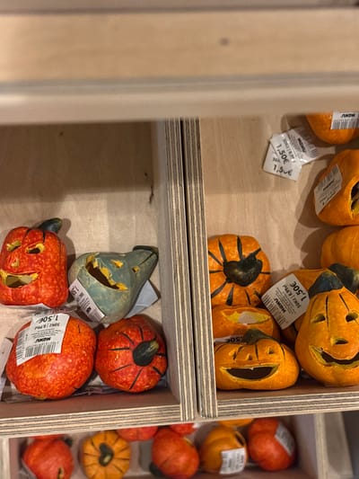 The scene, located in Capital City of Prague, Czechia, depicts a close-up view of multiple small, decorative, artificial pumpkins and gourds arranged within a multi-compartment retail display unit constructed from light-colored plywood. Subjects include various orange pumpkins, some carved with jack-o'-lantern faces and others featuring black painted ribs; speckled red pumpkins with black stems; and one distinct teal-green gourd, also carved with a face. Several items bear white price tags, clearly indicating "1,50€", alongside smaller text such as "MONU" and "50Kr / 6,99". No individuals are present; the composition focuses entirely on the merchandise displayed for sale.