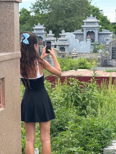 A young woman with long, dark, wavy hair, adorned with a light blue bow, stands in a graveyard in Hoa Lư Ward, Vietnam. She is viewed from behind, wearing a black sleeveless dress with a white trim and a white bralette. The woman is holding a smartphone in her right hand, angled to take a photograph. She is positioned next to a tan, rough-textured wall on the left side of the frame. In the background, numerous grey, ornate gravestones and structures, characteristic of Vietnamese cemeteries, are visible amidst lush green vegetation and trees under an overcast sky. The ground in the immediate vicinity of the gravestones appears to be paved with reddish-brown bricks.