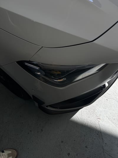The image provides an overhead perspective of the front-right quarter of a white Mercedes-Benz car, parked on a light grey, textured concrete surface. The car's pristine white bodywork, including the bonnet and front fender, is visible, featuring sharp panel gaps and subtle reflections of the environment.

The intricate right headlight is a prominent feature, displaying multiple LED elements. Faint, embossed text within the headlight assembly clearly reads "MULTIBEAM LED". Below the headlight, a dark, aerodynamic air intake or vent is integrated into the front bumper, next to which a small, circular parking sensor is visible. A portion of the car's dark wheel and tire is also discernible beneath the fender.

Strong, angular shadows fall across the concrete ground and partially obscure the lower front section of the car, indicating a direct light source from above, possibly daylight filtering into a garage, carport, or covered parking area. In the lower right corner of the frame, the tip of a person's right foot, wearing a light-colored sneaker (resembling an Adidas Samba with suede and white accents), is partially visible on the concrete. The scene is static, devoid of explicit activity, suggesting the car is parked. No specific location cues for Cape Town, South Africa, are visible within the image itself beyond a generic parking setting.