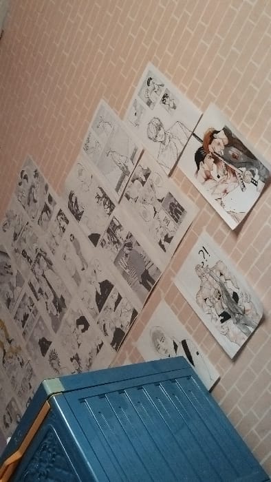 The image shows a wall covered with manga pages, featuring black and white illustrations of people and scenes. The wall is made of light pink brick-like tiles. A blue plastic storage container with a decorative lid is placed in the bottom right corner. The scene appears to be an indoor setting, possibly a bedroom. The manga panels depict various characters and storylines, likely from a single series or collection. The overall impression is one of an individual's personal space, decorated with a passion for manga. The location is a residential area in Panjalu, Indonesia, with coordinates -7.105 latitude and 108.272 longitude.