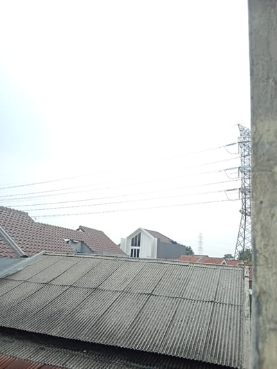 This media file captures a slightly overcast sky over a residential area in Depok, Indonesia. The foreground is dominated by the corrugated metal roof of a building, showing signs of age and wear. Behind it, several other rooftops are visible, including those with reddish-brown tiles and a modern white house with a distinctive gabled window.

In the background, a large electricity pylon stands tall, with multiple power lines stretching across the frame, indicating urban infrastructure. The sky is bright and diffused, suggesting it is daytime, likely morning or late afternoon, given the lack of harsh shadows. The overall scene is one of a typical suburban or urban landscape, with a focus on the interplay between human dwellings and the surrounding environment. There is no visible text within the image.