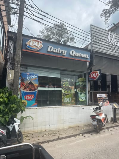 A Dairy Queen store is shown with its sign, "Dairy Queen DQ", prominently displayed. The store has window advertisements for Blizzard treats, including matcha and pistachio flavors. Thai script is present on a promotional banner. Scooters are parked outside. A nearby sign says "Narya's." Utility lines are visible in the foreground with an outdoor electrical box.