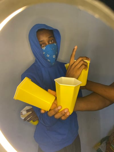 This image, captured in Ngleshi Amanfro, Ghana, displays a partially obscured individual in an indoor setting. A person wearing a blue hooded sweatshirt and a blue bandana covering the lower face, revealing only their eyes and forehead, is centered in the frame. This individual holds two yellow, hexagonal plastic cups in their left hand, and their right index finger is extended upwards. Another arm, entering the frame from the right, holds two more yellow cups. A distinct, bright, curved light source, consistent with a ring light, is visible in the upper and lower left corners. The background consists of a plain light gray wall, with a small segment of patterned fabric visible in the bottom right corner. The central individual's gaze is directed towards the viewer.