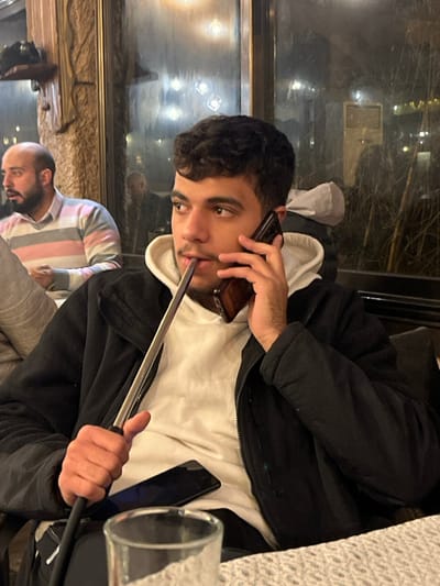 A young man in a black jacket over a white hoodie smokes a hookah while talking on a cell phone. He looks directly at the camera. His other hand holds the hookah hose. To his left, an older man sits with a striped shirt. A second phone and glass are on the table. A window with blurred city lights is visible in the background.