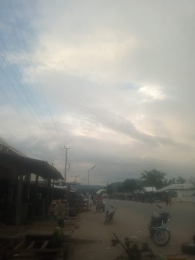 This image depicts a street scene in Serti, Nigeria. The time of day appears to be late afternoon or early evening, with the sky filled with clouds and a soft, diffused light. The weather seems overcast, possibly leading up to or following rain.
The scene is a dusty, unpaved street lined with small shops or market stalls on the left. These structures have corrugated metal roofs and open fronts, displaying various items for sale, including bottled drinks stacked on shelves. On the right side of the street, more buildings and shops are visible, along with people and motorcycles.
Several motorcycles are parked on the street, suggesting transportation is a common mode of travel in this area. One motorcycle in the foreground right is red and green, with a turquoise fender. Another, on the left, is white. People are present, though most are not clearly identifiable due to the image's quality. One person in the left foreground, wearing a green and yellow shirt, is seated on a wooden structure. Other figures are visible in the background, moving around or standing near the shops.
Power lines crisscross the sky, supported by utility poles, indicating the presence of electricity infrastructure. The overall atmosphere is that of a typical, busy market street in a Nigerian town, with a sense of everyday life unfolding. There is no visible text in the image.