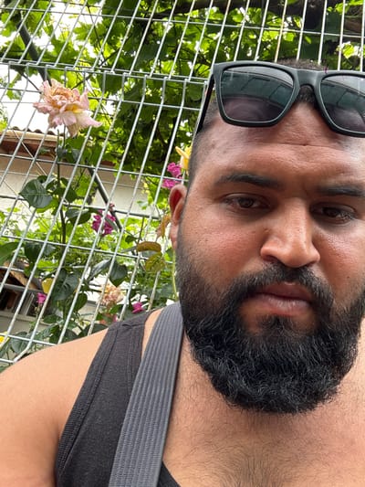 This close-up portrait, likely a selfie, features a man positioned outdoors. His head is slightly tilted, and he looks directly at the camera with a neutral expression. He has dark hair, a full dark beard, and a mustache. Dark sunglasses with dark lenses are pushed up onto his forehead. He is wearing a dark gray or black sleeveless top, with a lighter gray strap visible over his right shoulder.
The background is dominated by a light-colored metal wire grid, resembling a trellis or fence. Through and behind this grid, dense green foliage, including leaves and vines, is visible. Several flowers add color to the scene: a large, faded light pink and white rose appears in the lower left, while smaller vibrant pink flowers are scattered throughout the greenery. A faint architectural element, possibly a building, is discernible in the distant background. The scene is brightly lit by natural daylight, suggesting a clear day. This image was captured in Konstantinovo, Bulgaria. There is no visible text.