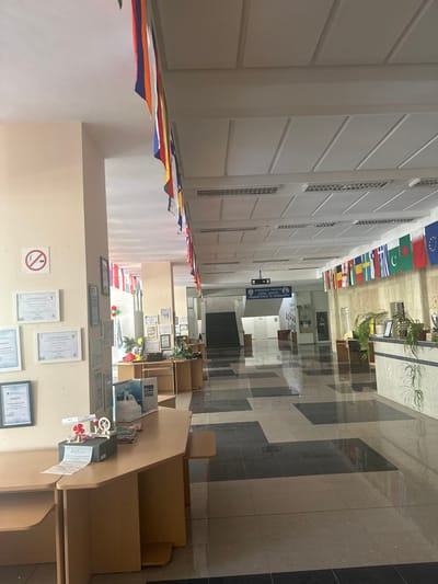 The image depicts the interior hallway of a building, likely a school or educational institution, located in Dobrich, Bulgaria. The hallway features a checkered tile floor with dark gray and light beige squares. The walls are painted a pale beige. Along the left wall, several framed certificates and a "no smoking" sign are displayed. A light brown wooden desk with various items, including books, a small model of a turbine, and printed papers, is positioned against the left wall.
The hallway stretches into the distance, with a staircase visible on the left side further down. On the right side of the hallway, a counter area is present, with plants in pots and framed items on display. A darker colored wall is visible behind the counter.
A prominent feature of the hallway is the series of national flags strung along the upper part of the walls on both sides, indicating an international or multicultural environment. The ceiling is white with a tiled panel design and ventilation grilles. The lighting appears to be fluorescent, integrated into the ceiling panels. The overall impression is that of a well-maintained, institutional interior space. No people are visible in the image.