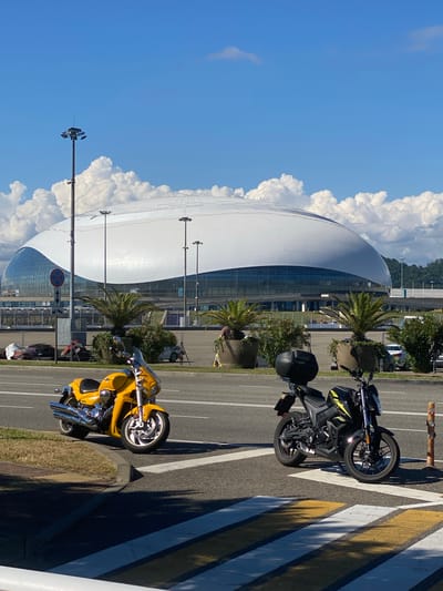 In the city of Sirius, Russia, a bright yellow cruiser motorcycle is parked on the side of a road, facing left, with its front wheel on a grassy embankment and its rear wheel on the road. Across the road, a black motorcycle with yellow accents is parked on a pedestrian crossing. In the background, a large, modern stadium with a white, undulating roof stands under a bright blue sky with scattered clouds. Palm trees and shrubs are planted in large planters between the road and the stadium. Streetlights are visible in the distance.